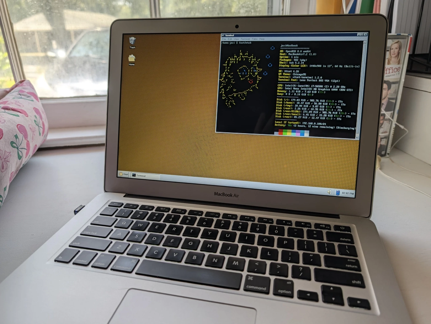 A photo of the BSDBook, sitting on my desk, with light shining behind it. The desktop is visible with a terminal emulator window open on it showing a fastfetch readout.
